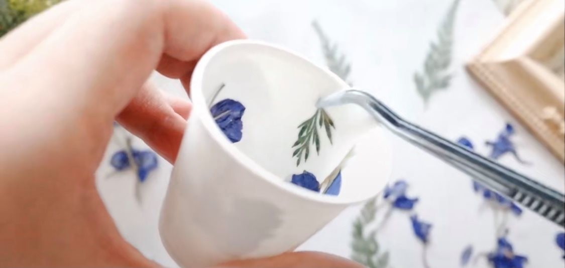 乾花蠟燭-1.第一步,首先把些少蠟溶掉,然後把乾花沾上一點點溶掉的蠟,為了牢固地把乾花黏在杯子上。然後,慢慢設計想要的花紋。最好用鉗子或夾子放進紙杯裏。