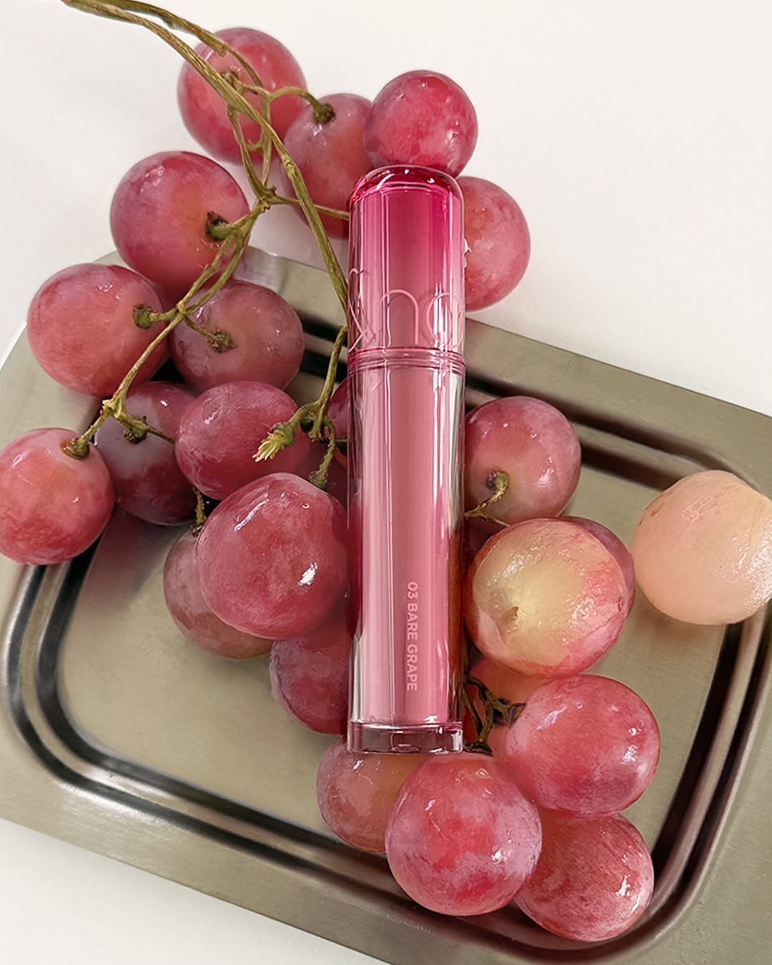 圖中展示的是Rom&nd Juicy Lasting Tint #03 Bare Grape，這款清透的冷調裸粉色能完美融合原生唇色，打造出自然不造作的「白開水」水光妝效。