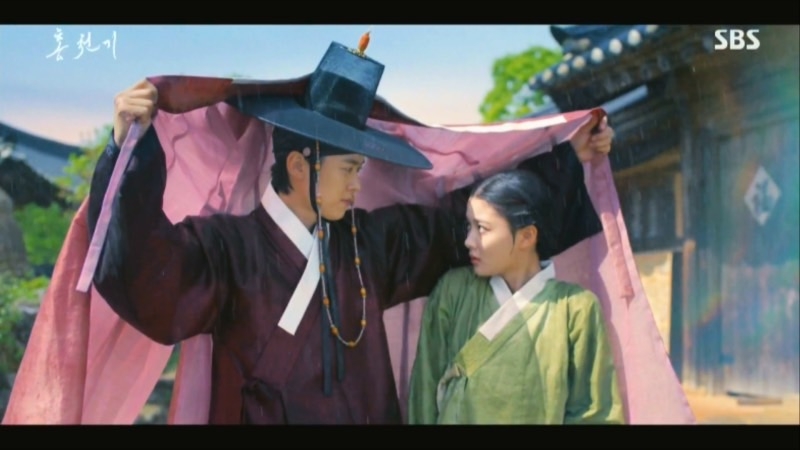 紅天機淋著雨走在街上，亮明大君忽然現身用衣服罩住她